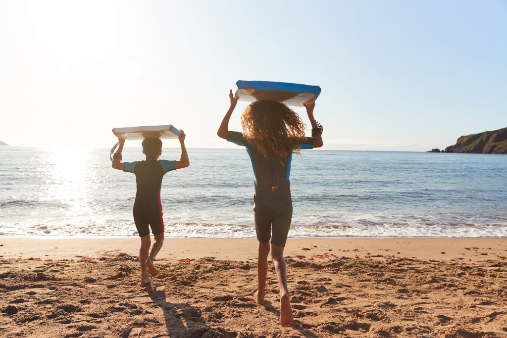 Rear View Of Children In Wetsuits Carrying Bodyboards On Summer Beach Vacation Having Fun By Sea