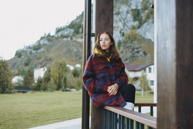 Young woman in coat sitting on wooden fence near cottage on the mountains background
