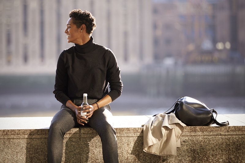 Young black woman wearing turtleneck sweater sitting in the sun on an embankment wall by the River Thames in London, backlit