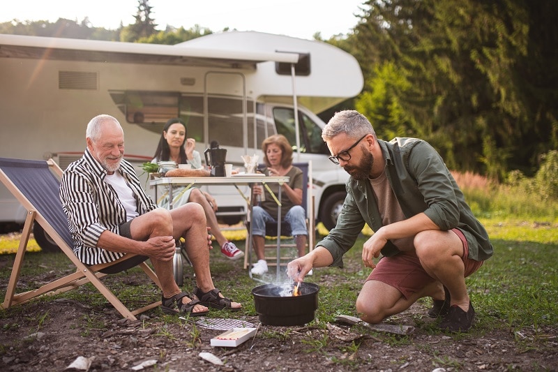 Multi-generation family preparing food on grill by car, caravan holiday trip.