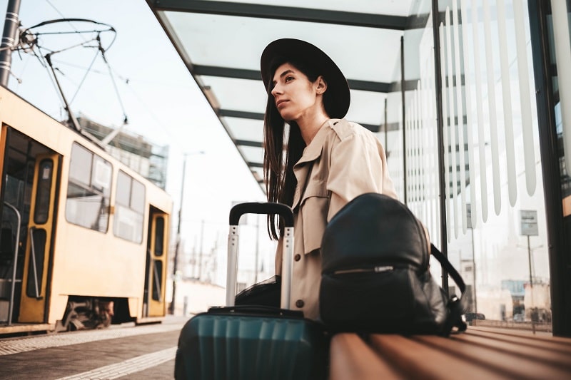 Young traveler lady in coat and hat with a sad face sits at a bus city stop alone