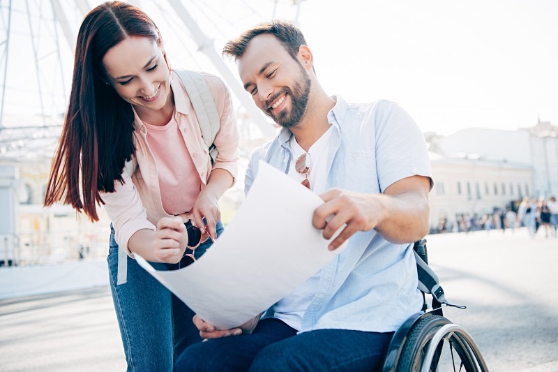 smiling handsome boyfriend in wheelchair and girlfriend looking at map on street