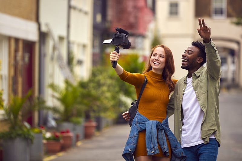 Young Couple Travelling Through City Together Vlogging To Video Camera On Handheld Tripod