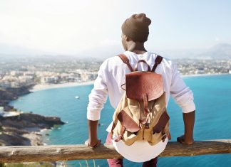 Travel for life concept. Outdoor shot of unrecognizable contemplating black tourist sitting at wooden beam of sightseeing platform admiring seaside view of resort city, carrying leather backpack