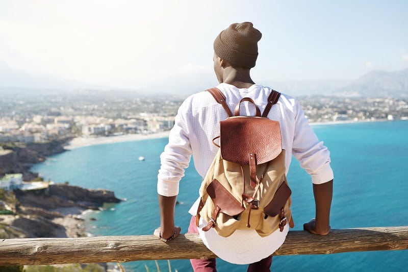 Travel for life concept. Outdoor shot of unrecognizable contemplating black tourist sitting at wooden beam of sightseeing platform admiring seaside view of resort city, carrying leather backpack